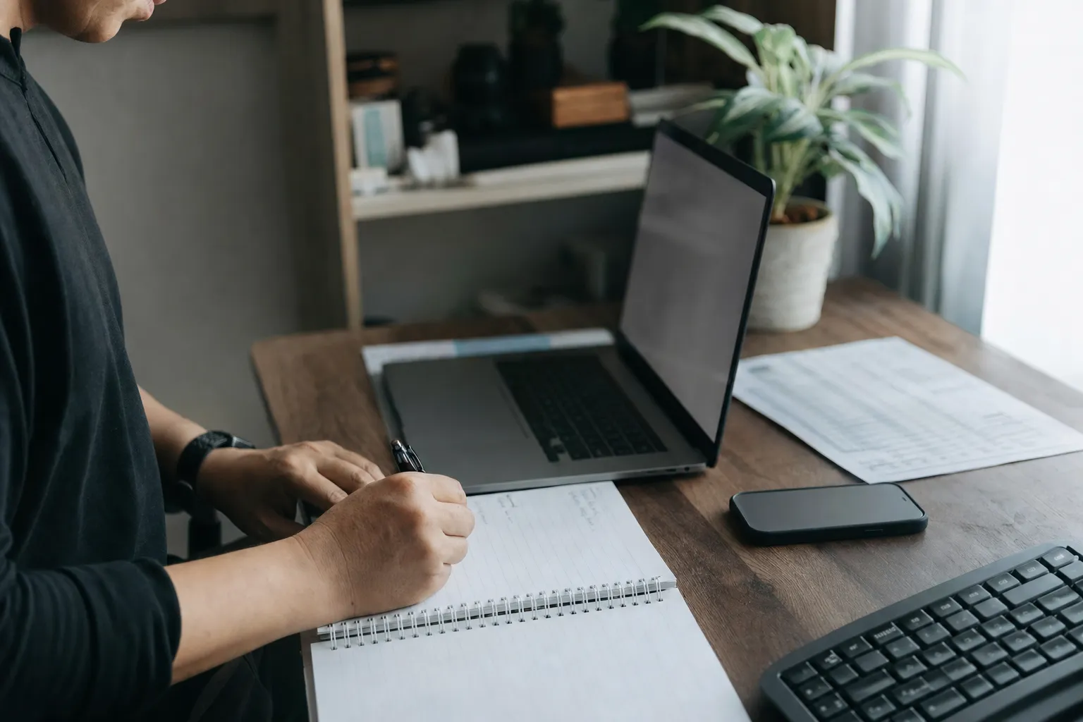A small home office with a laptop and a face-down statement, close on hands and notebook